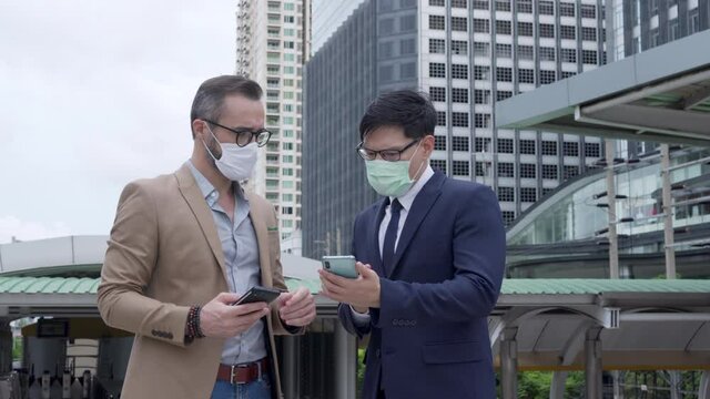Business people Caucasian with Asian man office worker colleague partnership wearing protective face mask for protect covid-19 virus discussing business project plan with using smartphone in the city.