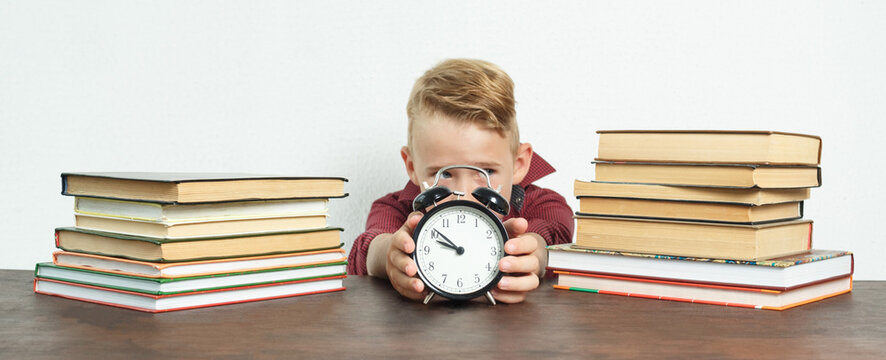 The schoolboy sits at the table and holds an alarm clock in his hands. Sharpness of the image on the alarm clock