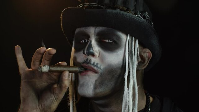 Creepy man with skeleton makeup in top-hat. Guy smoking cigar, making faces, looking at camera