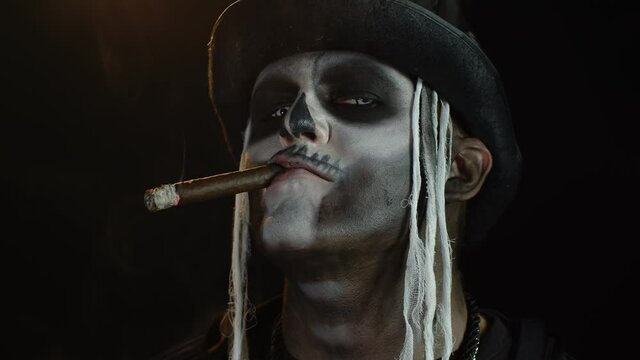 Scary guy in professional carnival makeup of Halloween skeleton smoking cigar, making faces, smiling