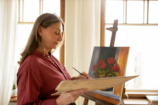 Portrait of talented beautiful middle aged female attending art workshop standing in front of easel, painting still life depicting flower pot. People, age, retirement, leisure and hobby concept