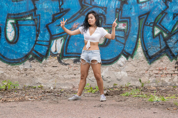 Fototapeta na wymiar A cheerful young and dark-skinned girl in a white top and blue shorts stands against the background of a wall with graffiti with her arms spread apart