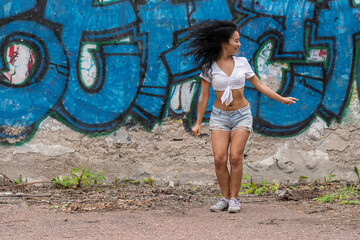 Fototapeta na wymiar A cheerful young and dark-skinned girl in a white top and blue shorts is dancing against the background of a wall with graffiti and scatters black hair to the sides