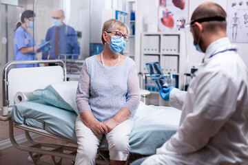 Doctor looking at x-ray of senior patient in hospital room during examination wearing face mask as...