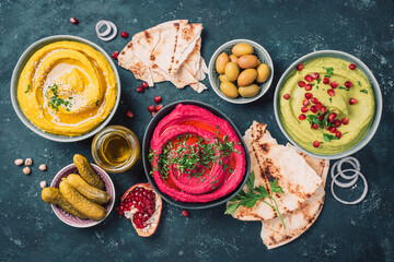 Ingredients for yellow turmeric hummus, green parsley hummus and red beetroot hummus on dark background, lemon, sesame, olive oil, pita, avocado, pomegranate. Middle eastern, jewish, arabic cuisine.