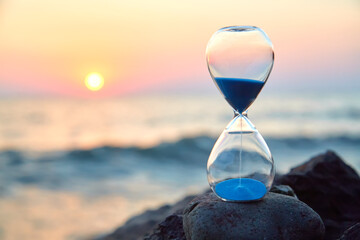 Hourglass with sand standing on rock. Sunset over sea and nature landscape. Running of time and...