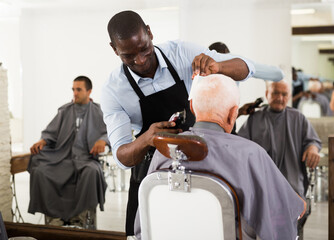 Professional hairdresser doing styling of elderly man with electric hair clipper at barber shop