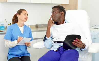 Obraz premium Male client with mirror having conversation with cosmetologist before procedure in clinic of esthetic cosmetology