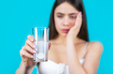 Woman taking drugs to releave headache. Brunette take some pills, holds glass of water, isolated on blue. Young woman taking pill against headache. Brunette taking a pill with a glass of water