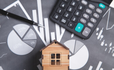 Wooden house model with a calculator and pen on financial graphs.