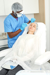 Cosmetologist man in mask preparing woman client for mesotherapy procedure in medical office