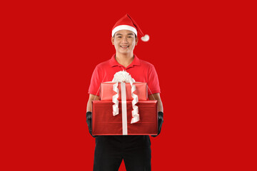 Happy young Asian delivery man in red uniform, christmas hat carry boxes of presents in hands on red background during christmas festivities