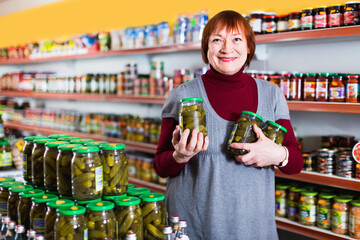 Adult cheerful smiling female customer holding glass jar of cucumbers in the supermarket