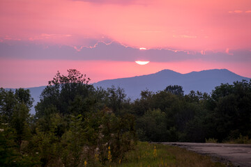 the sun rises against the background of the mountains. Green grass and a road stretching into the distance. Stanitsa Dakhovskaya, Republic of Adygea, Russia.