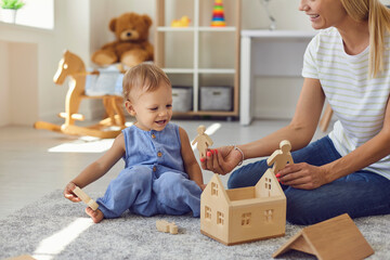 Joyful young nanny and little kid playing with wooden blocks in cozy nursery room - obrazy, fototapety, plakaty