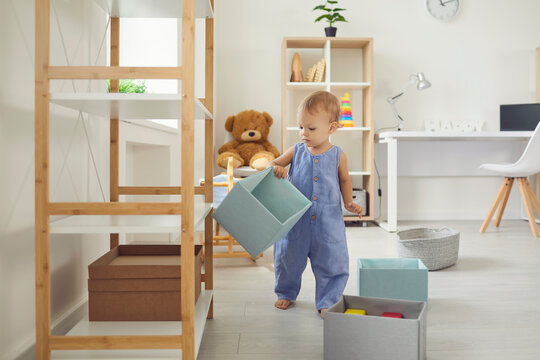 Cute Baby In Jumpsuit Standing At Home With Empty Box In Hand