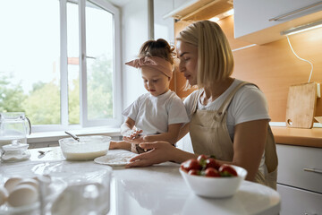 Obraz na płótnie Canvas happy mother baking with little daughter in apron and cook hat working with flour , bowl and spoon preparing dough teaching the kid baking and having fun together.