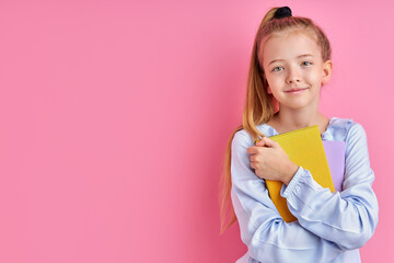 shy and diligent child girl is going to school isolated on pink background, she hold books and notebooks in hands, looking at camera and smiling