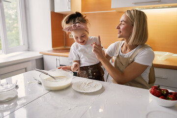 Obraz na płótnie Canvas happy mother baking with little daughter in apron and cook hat working with flour , bowl and spoon preparing dough teaching the kid baking and having fun together.
