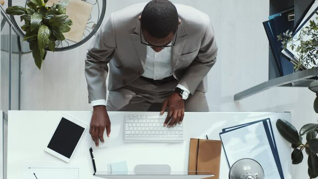 Top down shot of African American businessman in formal suit typing on wireless keyboard and using computer mouse while working at desk in the office