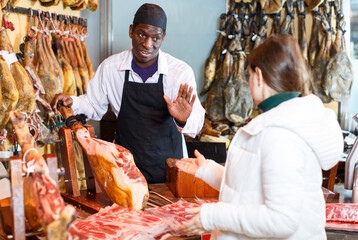 Happy successful butcher offering delicious Spanish ham to female client in meat delicatessen shop
