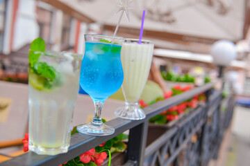 Three fresh juice drinks in tall glasses with plastic straws outside. Milkshake, blue curacao cocktail and mohito.