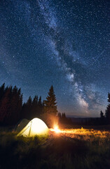 Burning campfire and illuminated tent under bright starry sky on which the Milky Way is clearly...