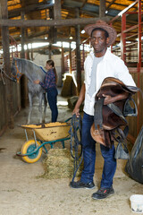 Farm workers man and woman preparing horse harness at stable