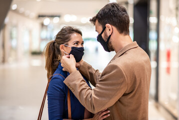 Attractive fashionable man adjusting face mask on his girlfriend while standing in shopping mall during corona virus.