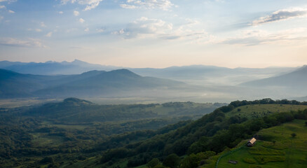 Fototapeta premium Early morning in the mountains. Morning sun and haze in the valley against the background of mountains. Mountain summer landscape. Small house in the mountains