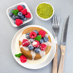Matcha pancakes served with condensed milk, blueberry and raspberry on white plate, top view, square format