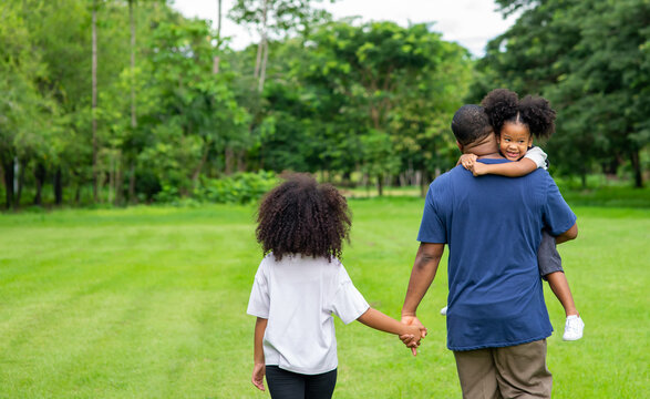 Happy affectionate mixed race family. African father carrying and holding hands his two little daughter walking in the park. Dad and cute child girl spending time together in outdoor weekend vacation.