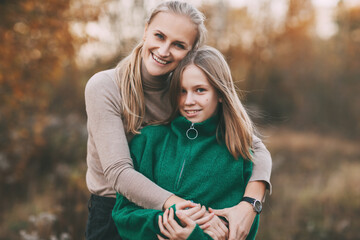 Fototapeta premium Portrait of happy mature mom and her blonde little daughter hugging and laughing while walking in autumn park outdoors.
