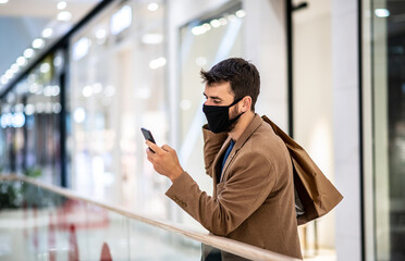 Young handsome fashionable man with protective face mask on standing in shopping mall, carrying bags and using phone during corona virus outbreak.