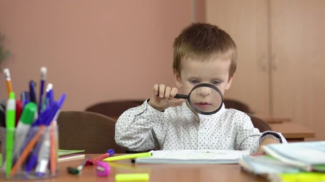 A handsome three-year-old boy sits at a school desk and looks at a notebook through a magnifying glass. Preschool education. 4k