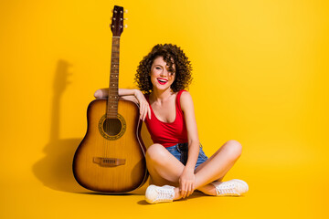 Portrait photo of young girl musician with curly hair holding keeping hand touching guitar instrument sitting down isolated on vivid yellow color background - obrazy, fototapety, plakaty