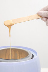 Wax heater with hot yellow wax on wooden spatula in human hand. Hot wax for depilation. Salon...