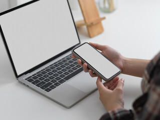 Young female holding mock up smartphone while using mock up laptop computer