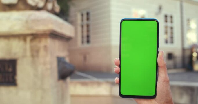 Close up view of female hands with trendy fingernails design holding and pressing on phone with mockup screen. Old city fountain at background. Concept of greenscreen and chroma key.