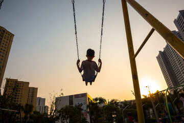 Child swinging on swing in sunset in city with building on background - obrazy, fototapety, plakaty