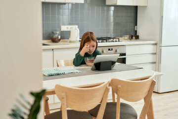 A little girl is watching a tablet attentively while sitting alone at a table and eating her quick breakfast with a spoon in a bright large kitchen at home