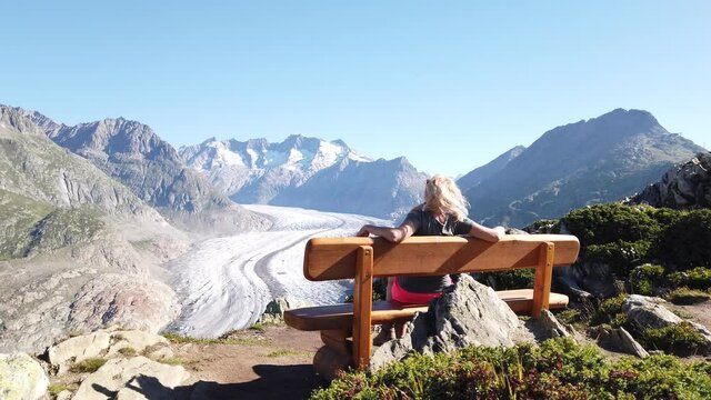 Aletsch Glacier from Moosfluh viewpoint in summer with clear blue sky in Swiss Alps, Valais Canton, Switzerland, Europe. Tourist woman relaxing sitting in a wooden bench looking over glacier.