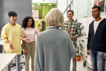 Cheerful young team greeting new employee, Back view of aged woman, senior intern waving at her colleagues in the modern office