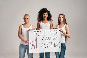 Three confident diverse women in white shirt and casual denim jeans holding, standing with a banner in their hands isolated over grey background