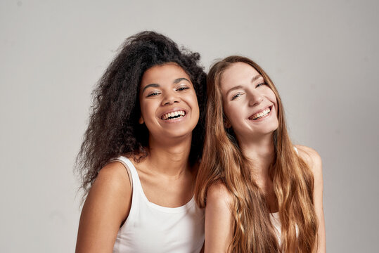 Portrait of two happy young diverse women in white shirts smiling at camera while posing together isolated over grey background