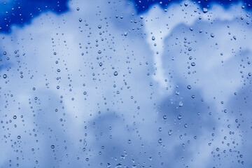 blue rain water drops on a window glass close up with blue colorful clouds in background, drop and cloud macro texture ; bad weather backdrop