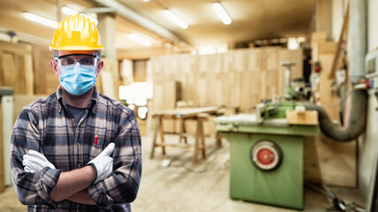 Carpenter worker in the carpentry workshop, wears helmet, goggles, leather gloves and surgical mask to prevent coronavirus infection. Preventing Pandemic Covid-19 at the workplace.