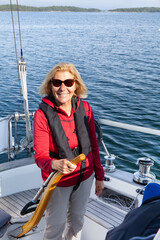 Happy blonde senior woman with sunglasses steering her sailboat out on the sea wearing a life jacket