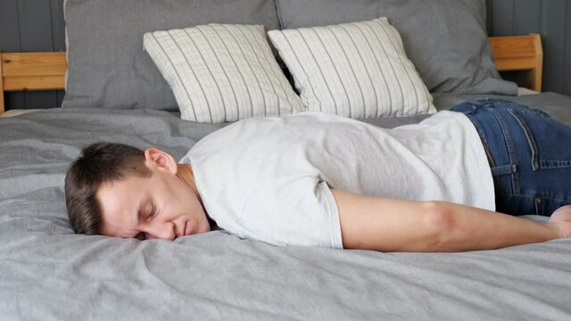 Tired young guy in t-shirt falls down on belly on large bed with grey linen and designer pillows in contemporary room at home closeup