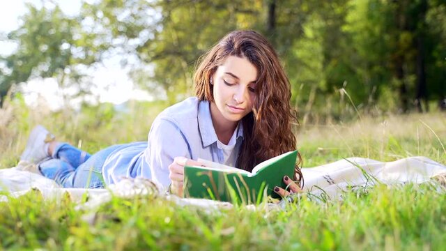 Beautiful young lady with curly dark hair lying on soft blanket and reading interesting book at summer park. Cute female relaxing on fresh air with new literature.